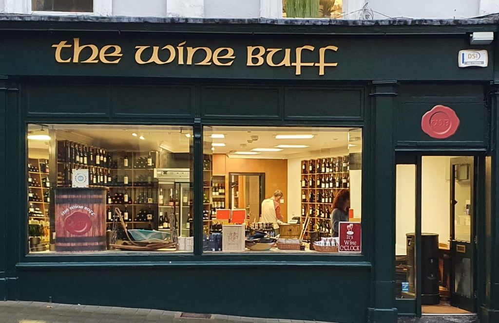 The Wine Buff shop front with green coloured walls and 2 large windows with wine displayed in the windows and the shop lit up with bright light and wines stacked on shelves. The Wine Buff logo over the shop in gold text.