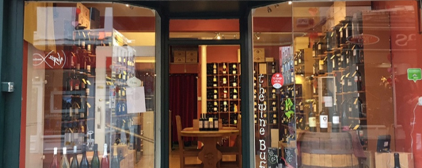 A picture of The Wine Buff Cork with a door between the two windows, the shop has green colour frontage, inside you can see wine shelves, wine barrels and wine bottles.