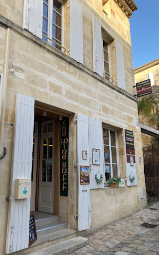 Picture of The Wine Buff shop in St Emilion, France. The facade of the shop is in large stonework yellow sandstone colour, in french style. There are grey shutters on the windows and doors with a terrace seating area on the right.